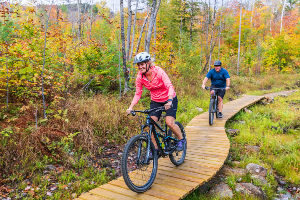 A couple mountain biking over a boardwalk on a trail in the Bethel Community Forest in Bethel, Maine.